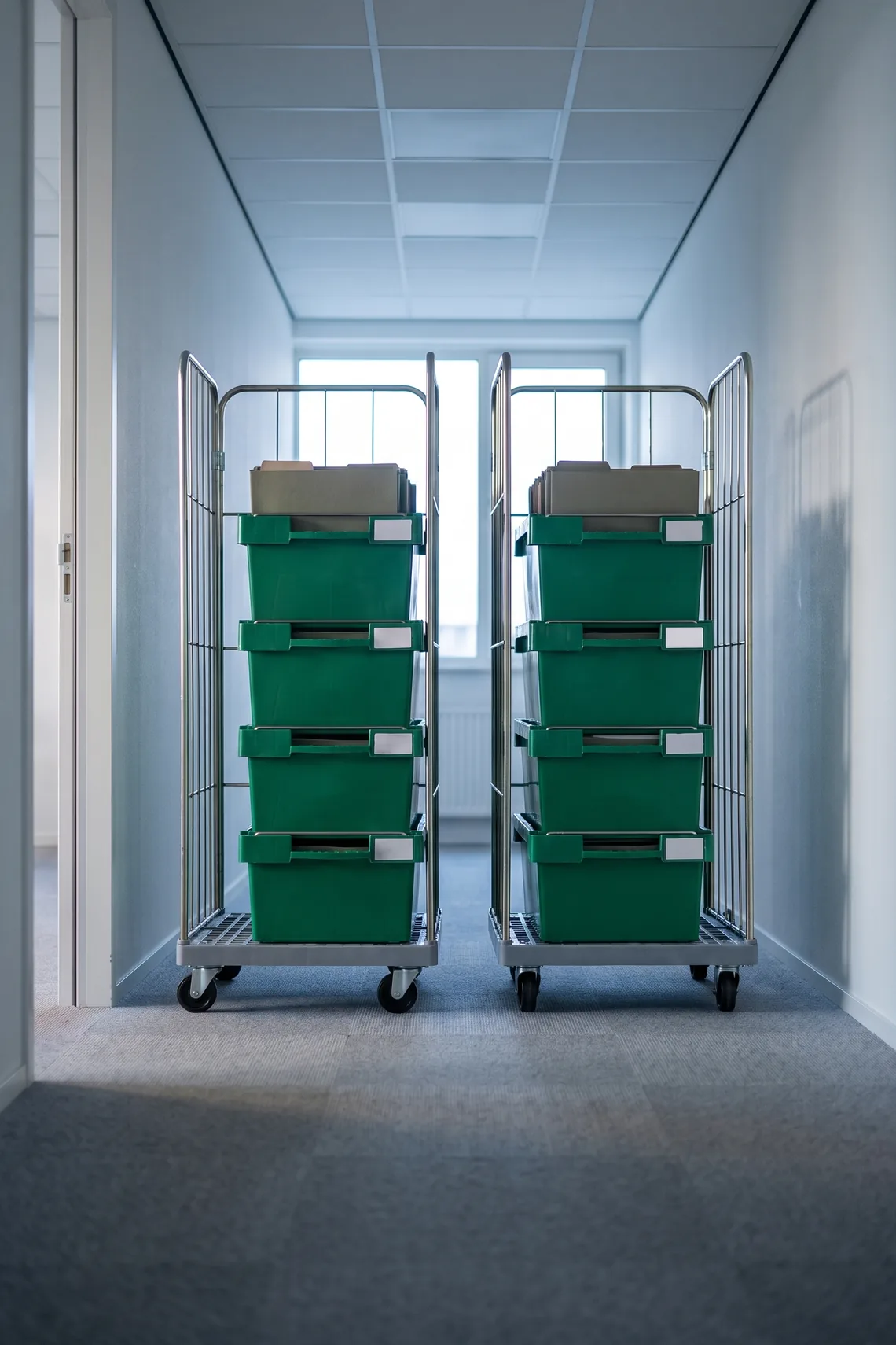 Twee rolcontainers met groene kratten in de gang van een kantoor.