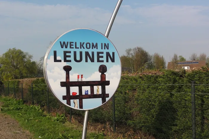 Sculptuur Welkom in Leunen: een rond bord aan een gebogen metalen paal, langs een haag
